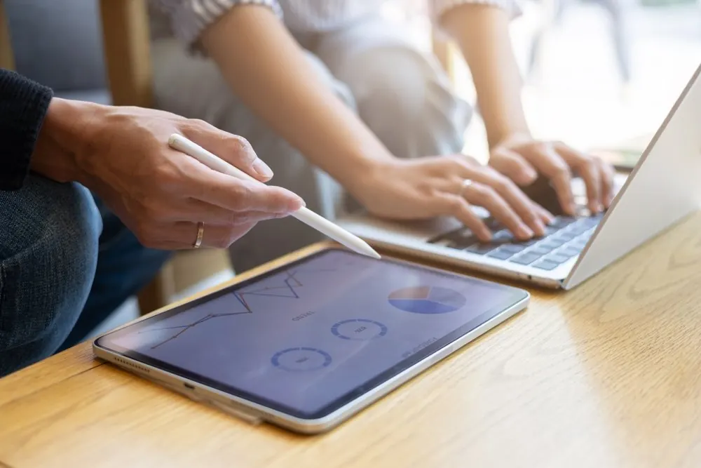 Hands on a tablet showing charts beside a laptop
